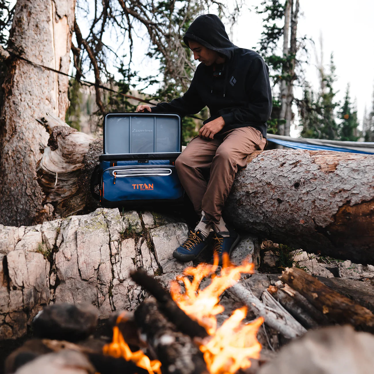 Person sitting by a campfire with a Titan cooler box in a forest setting