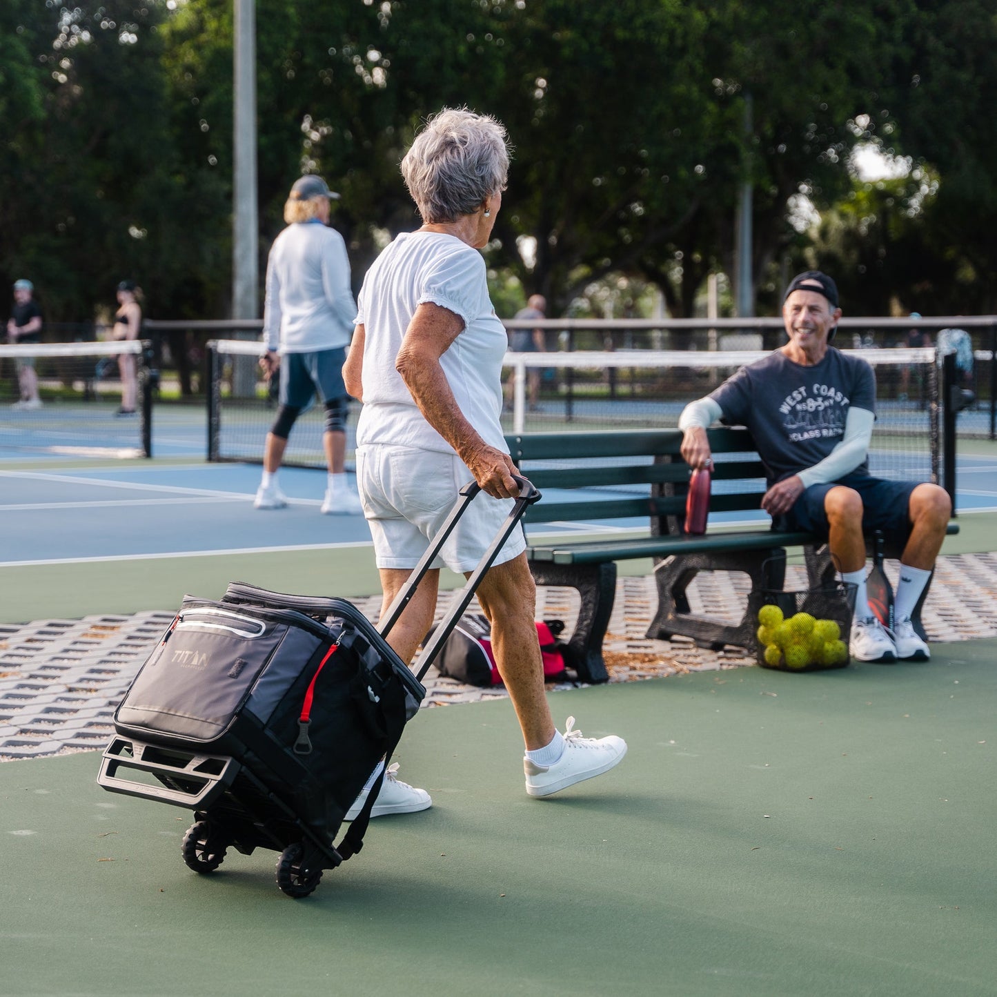 Person with a Titan wheeled cart on a tennis court with others in the background