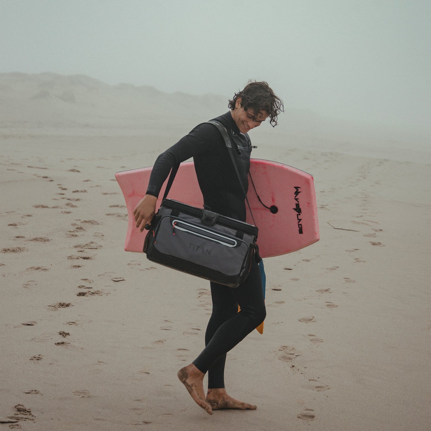 Person in a wetsuit holding a pink surfboard and a black Titan cooler on a sandy beach.